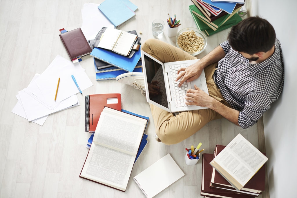 Young man sitting on the floor using laptop surrounded with great number of different things